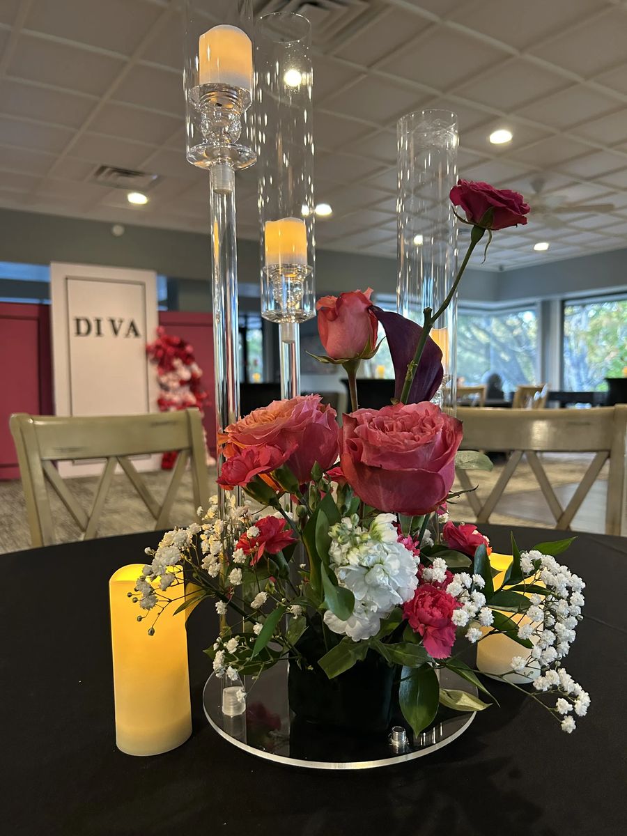 A rose and calla lily centerpiece with pillar candles and hurricane vases on a black tabletop.